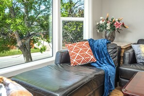 A room with a leather sofa, a patterned pillow, a vase with flowers, and a view of a tree through a window. - I @Home Entire 4BR3BA House/YYC/Parking/BBQ (Calgary)