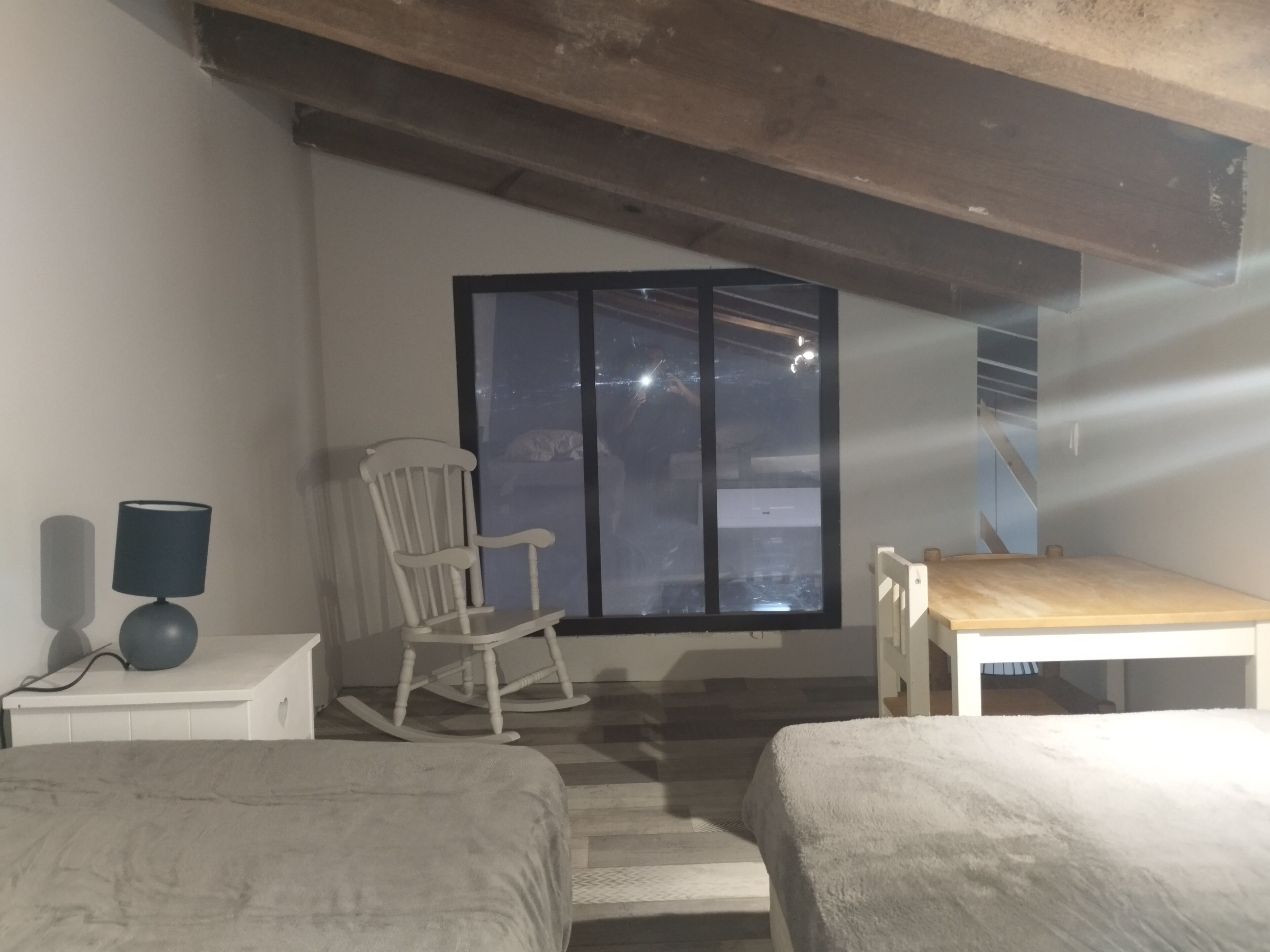 A bedroom with a wooden rocking chair, a small table, and a window with a view of the outside.