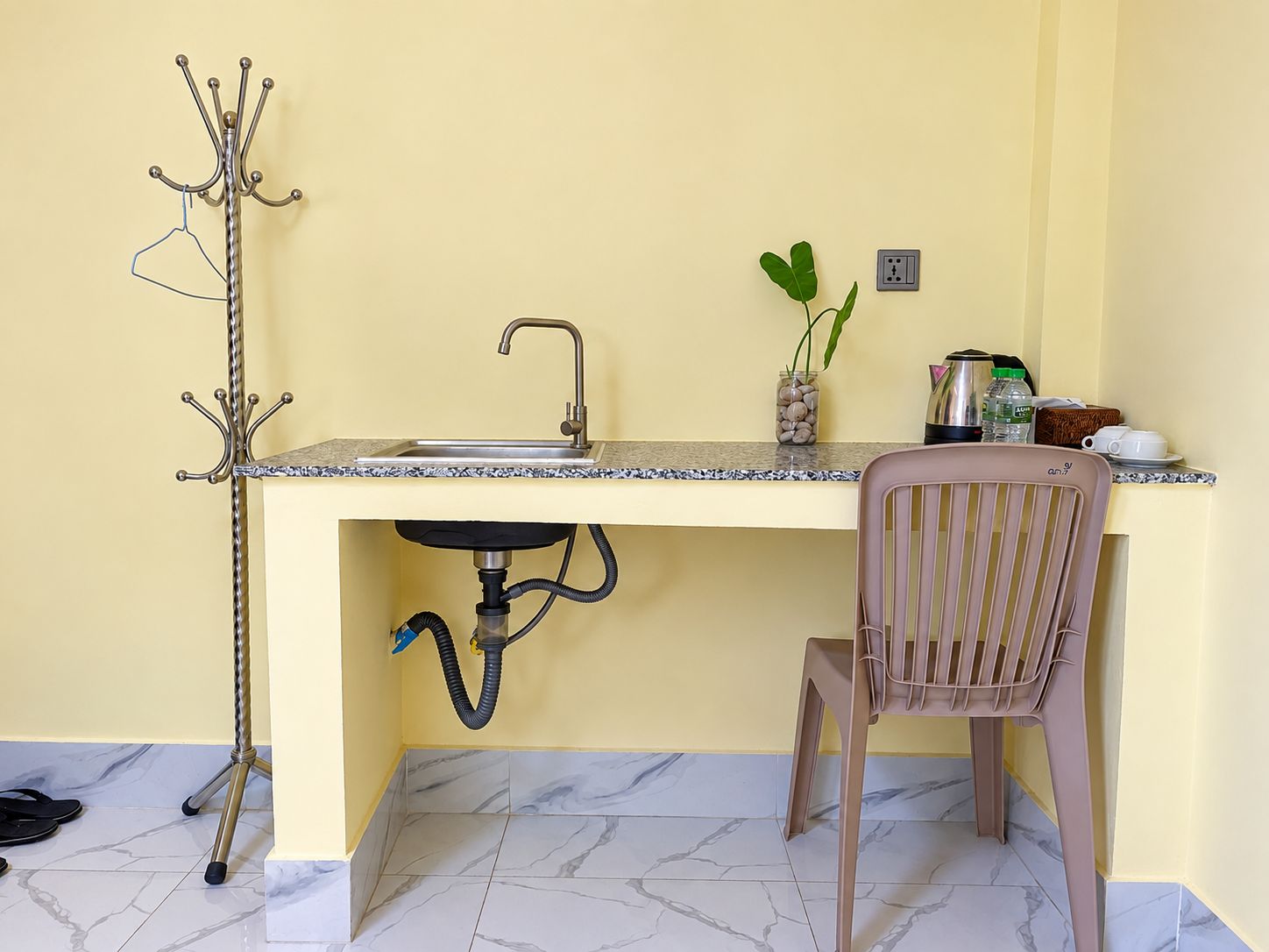 A compact kitchen with a sink, a chair, a coat rack, and a small plant on the counter.