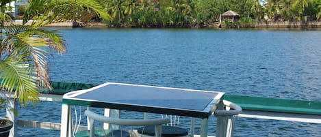 Una mesa y sillas en un barco, con vista a un cuerpo de agua tranquilo y palmeras al fondo.