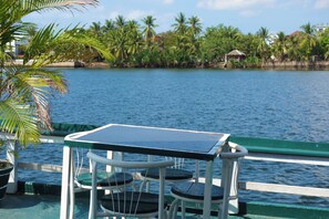A table and chairs on a boat with a view of a calm body of water and palm trees in the background. - AHHA River Breeze Kampot  (Kampot)