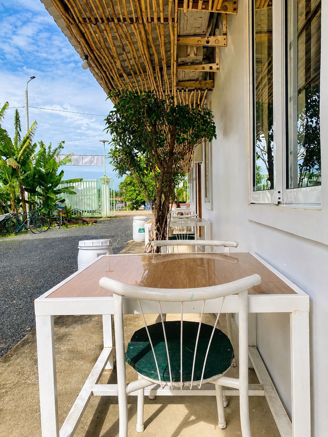 A table and chair set outside a building with a bamboo roof, a tree, and a view of the street.