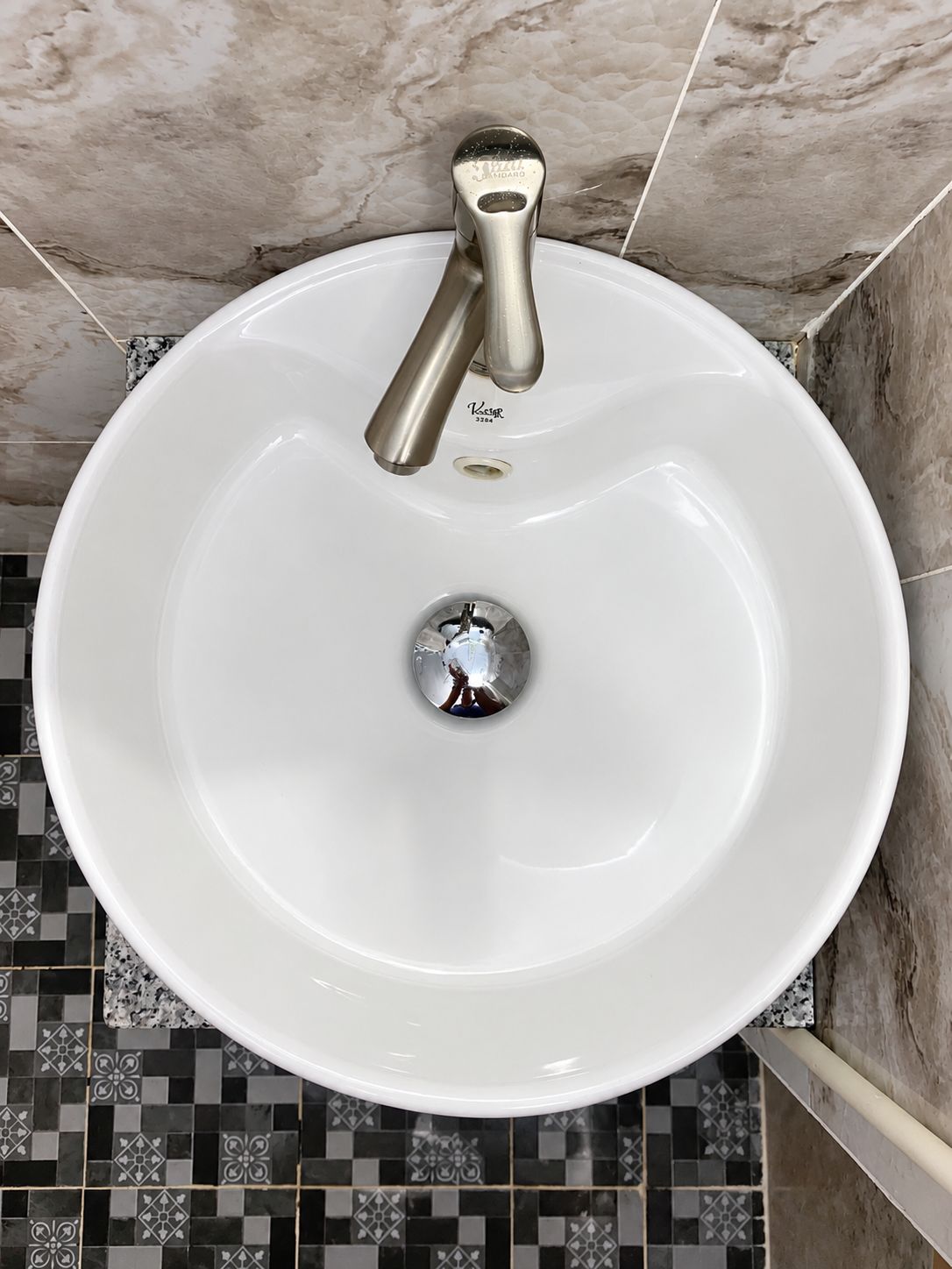 Un lavamanos blanco con un solo grifo, sobre un piso de baldosas con diseño geométrico.