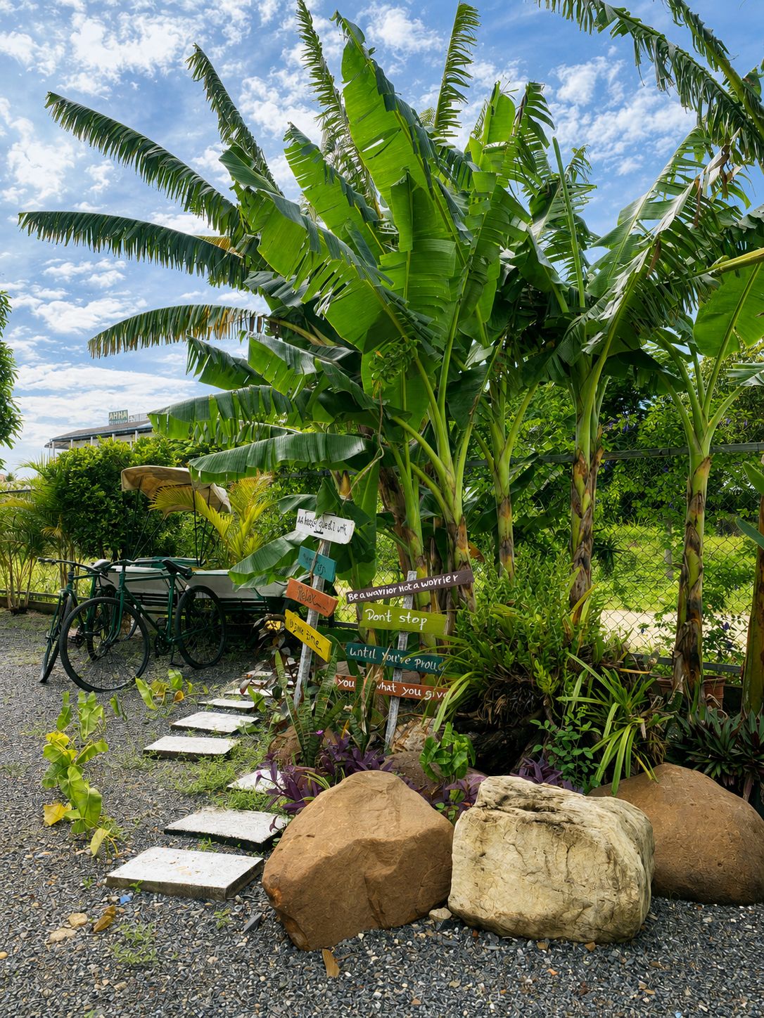 A garden with banana trees, a signpost with motivational phrases, a bicycle, and a paved path.