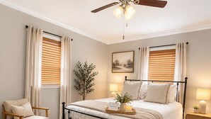 Ein Schlafzimmer mit Bett, Stuhl, Nachttisch, Stehlampe, Deckenventilator und einem Fenster mit Jalousien.