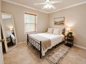 Un dormitorio con cama, mesita de noche con lámpara, ventana con persianas, ventilador de techo, puerta y alfombra.