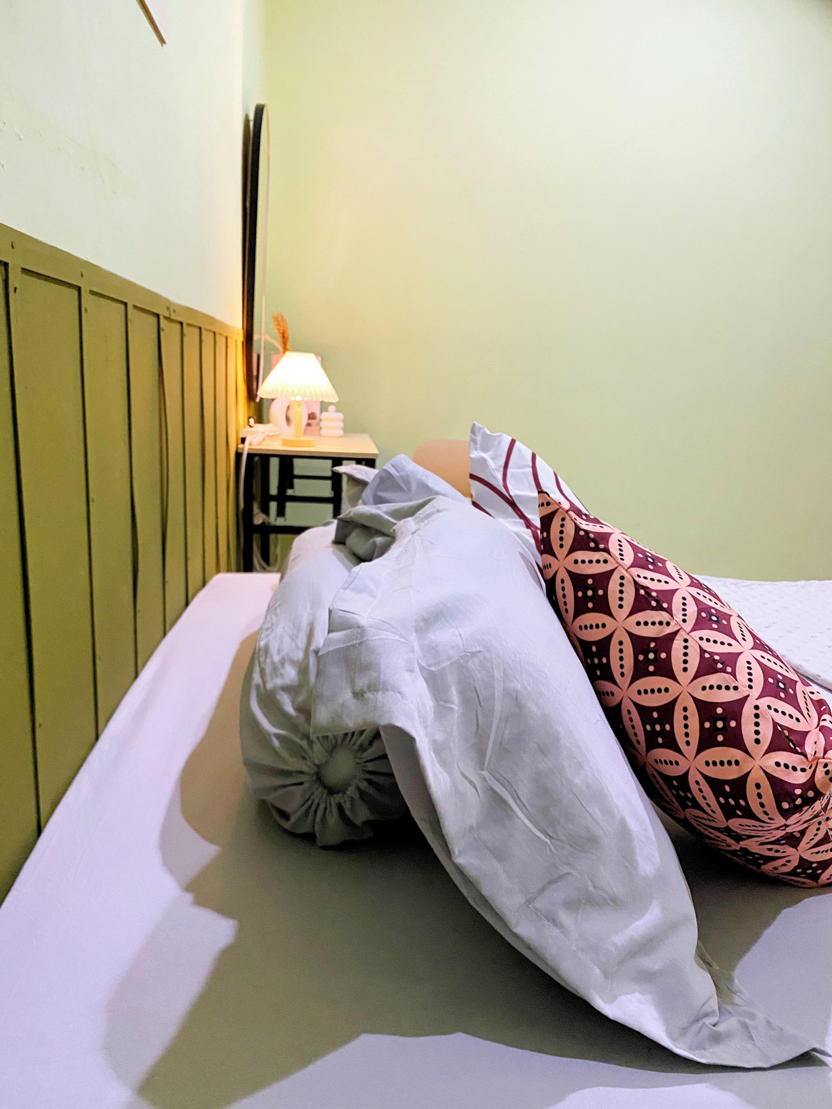 A bed with a white sheet, a patterned pillow, and a bedside table with a lamp.