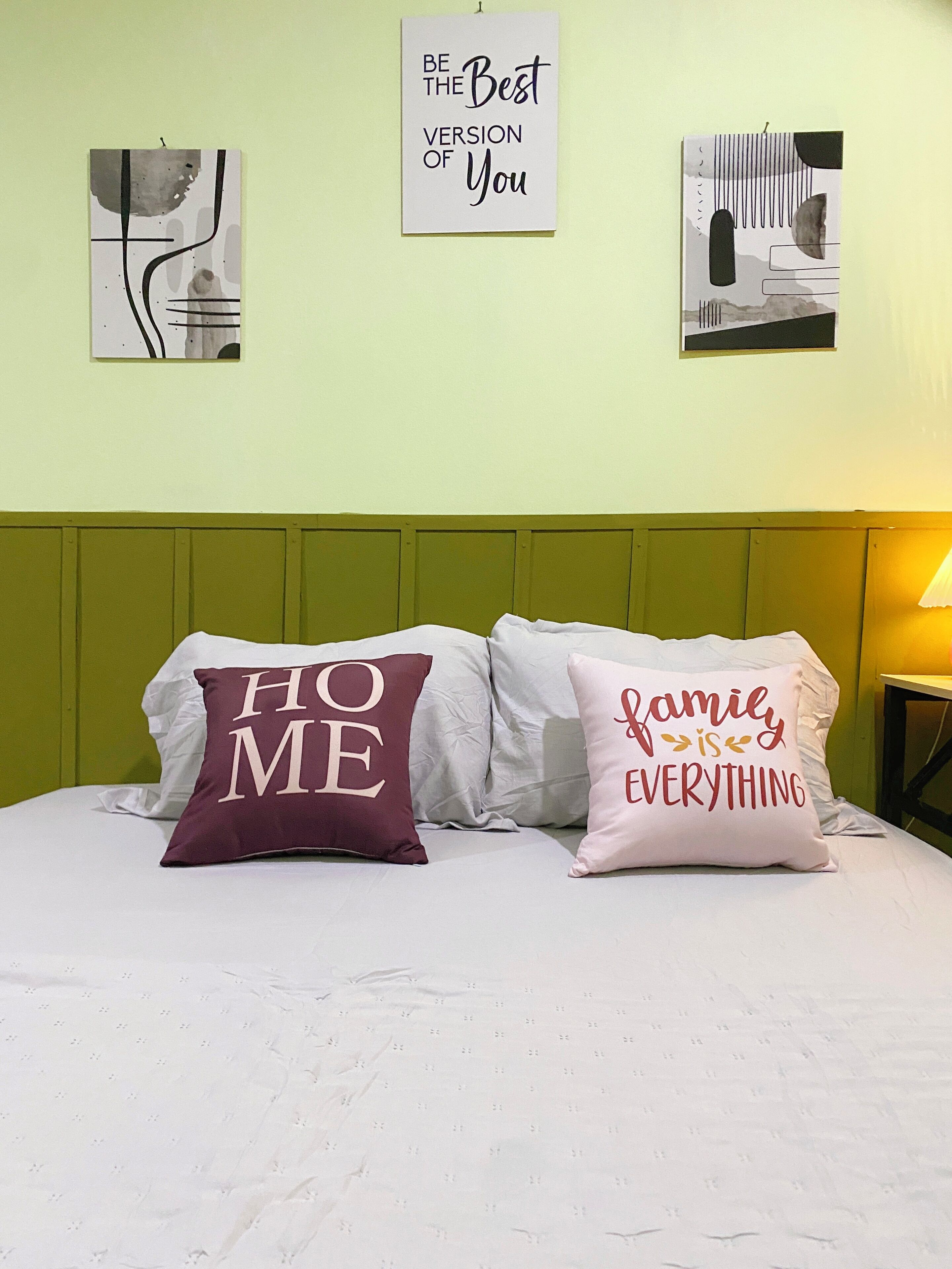 A bed with two pillows, one reading "HOME" and the other "family is everything," against a light green wall with two framed pictures.