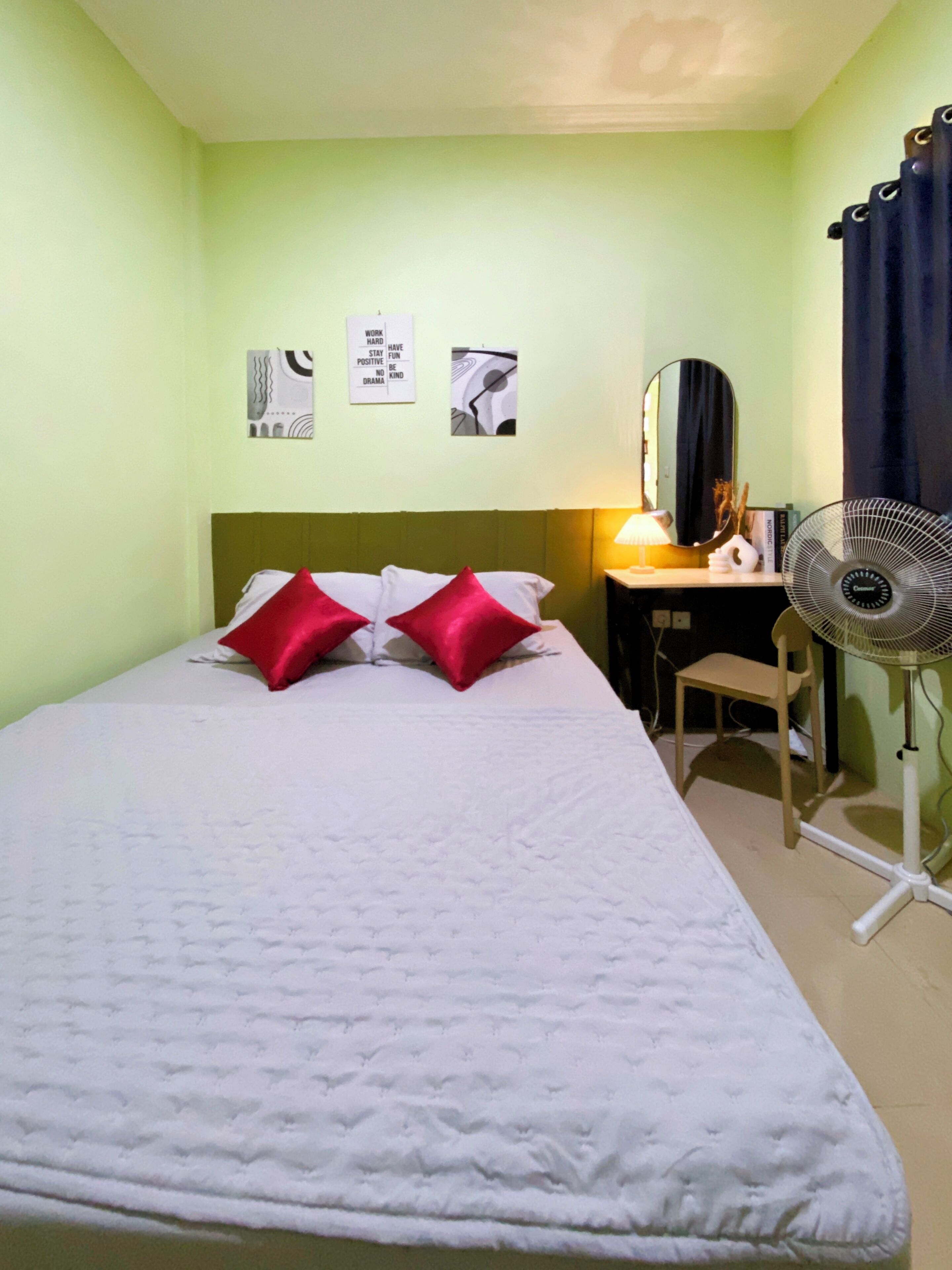 Une chambre avec un lit simple, un mur vert, un bureau avec une chaise, un ventilateur sur pied et deux tableaux accrochés au mur.