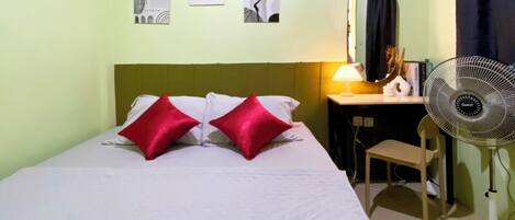 Habitación individual con una pared verde, un escritorio con una silla, un ventilador de pie y dos cuadros colgados en la pared.
