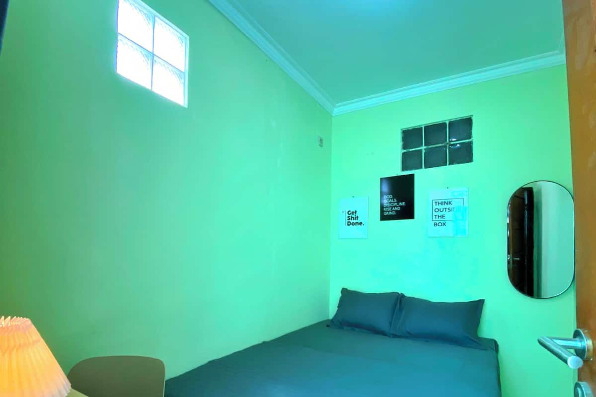 A small, brightly lit room with a single bed, a desk with a lamp, and a mirror on the wall.