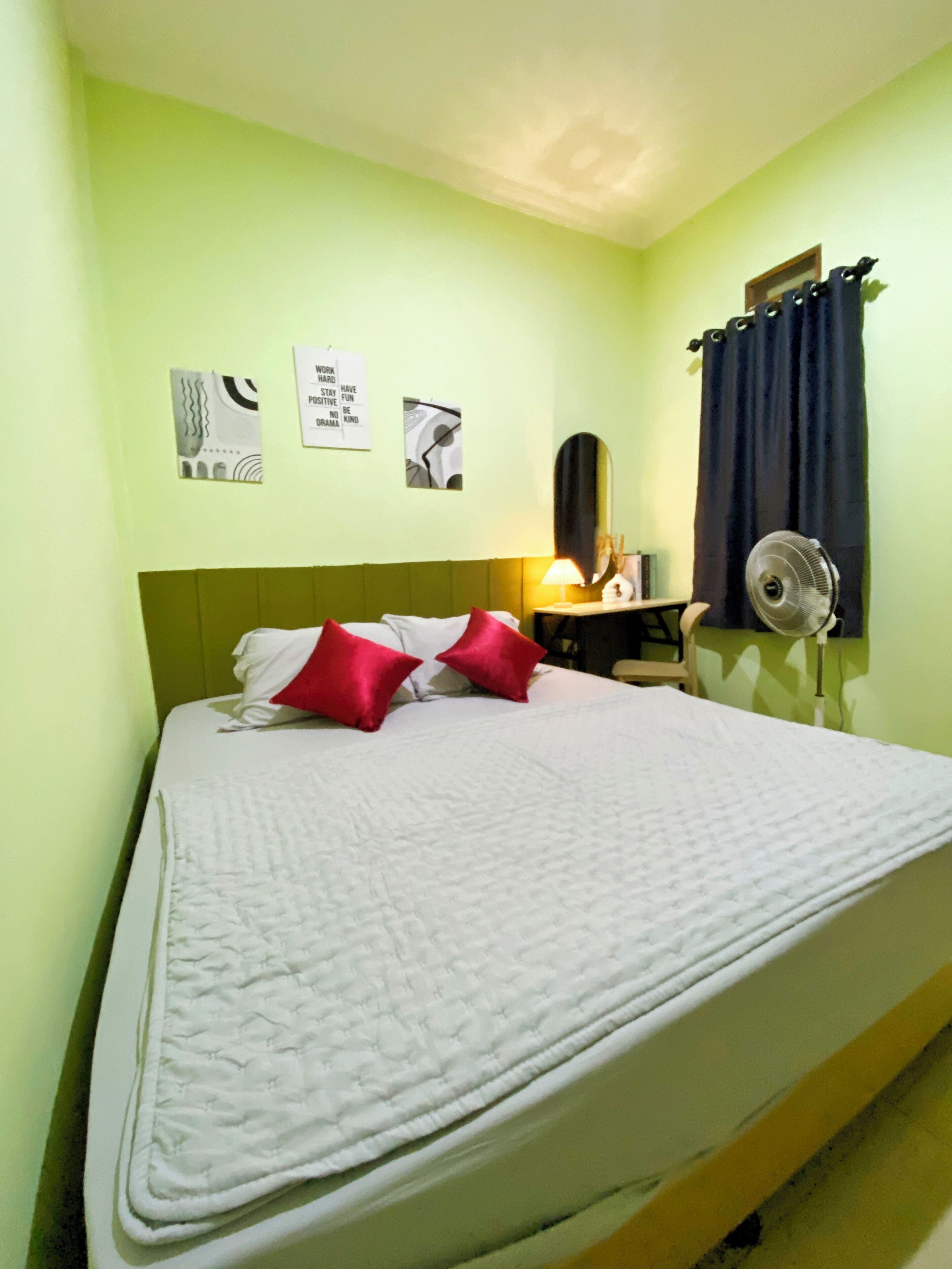 Una cama bien hecha con almohadas blancas y rojas, una lámpara de noche y un ventilador de pared.