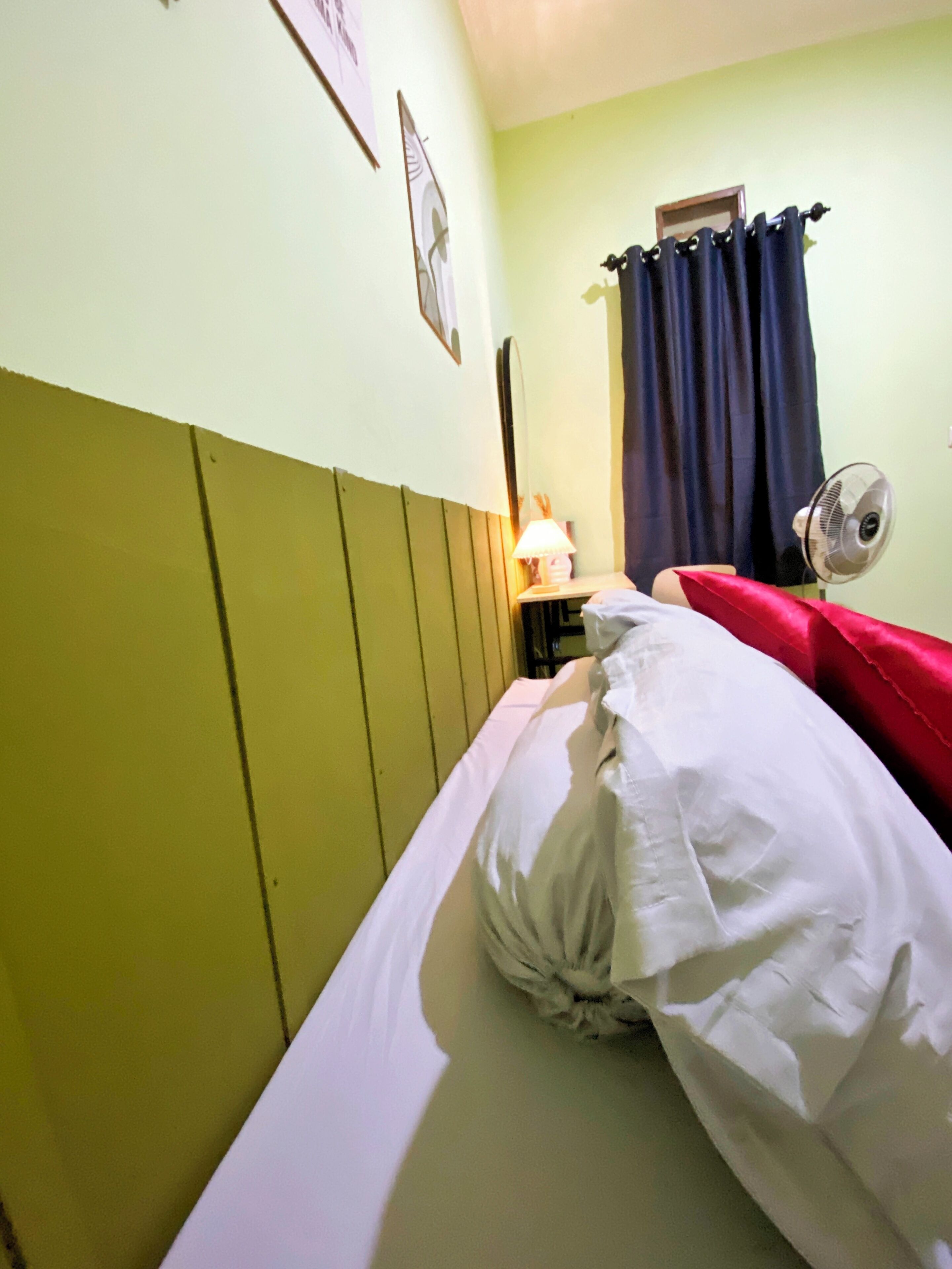 A single bed with white bedding in a room with green walls and a wooden headboard. There is a bedside table with a lamp and a fan.
