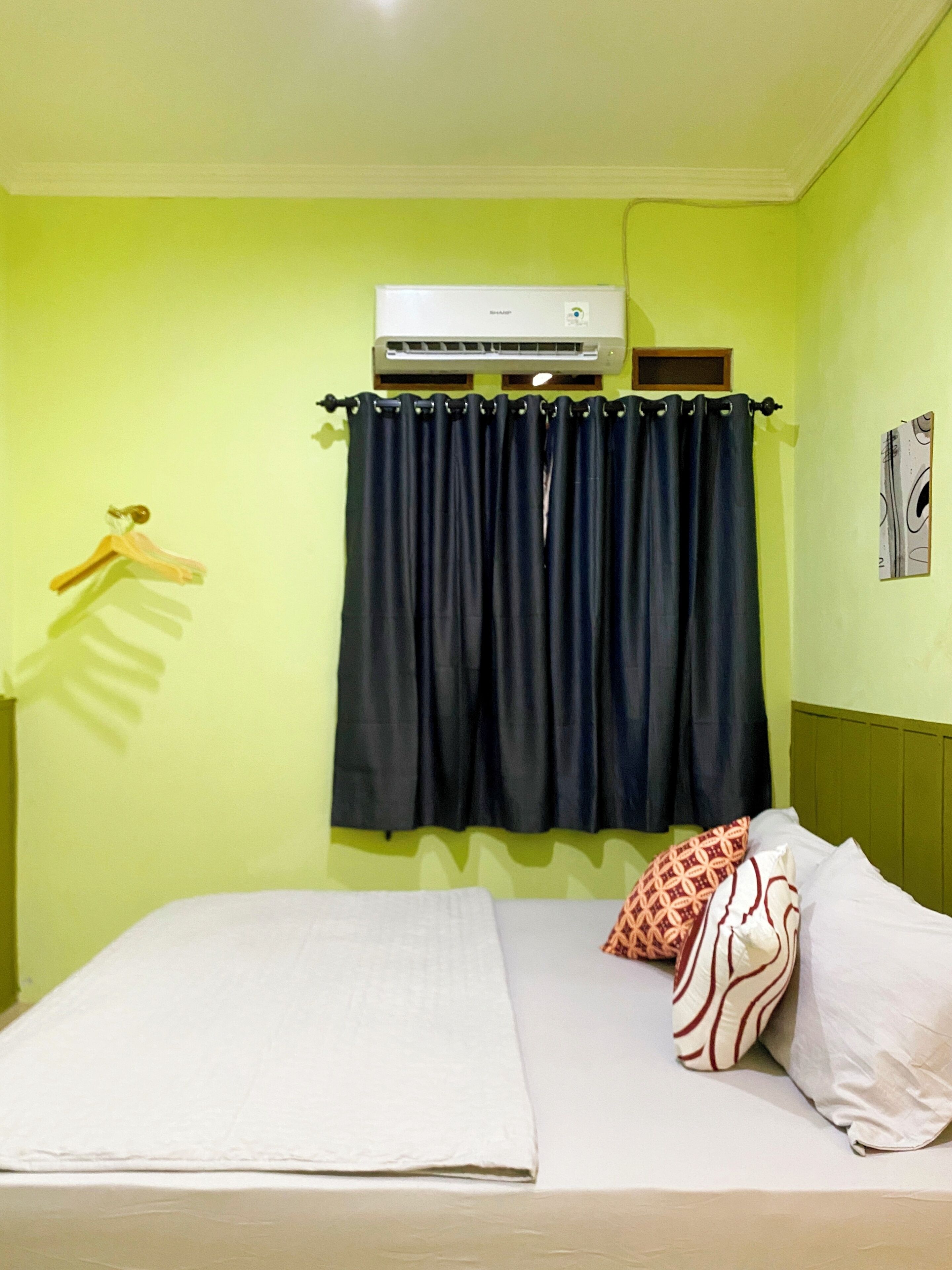 A hotel room with a bed, a hanging air conditioner, a dark curtain, and a wall-mounted picture.