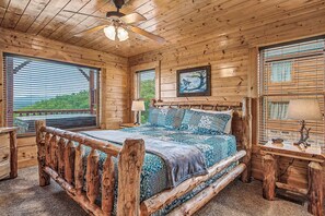 Ein Schlafzimmer in einer Blockhütte mit Bett, Holz-Kopfbrett, Deckenventilator und einem Fenster mit Jalousien.