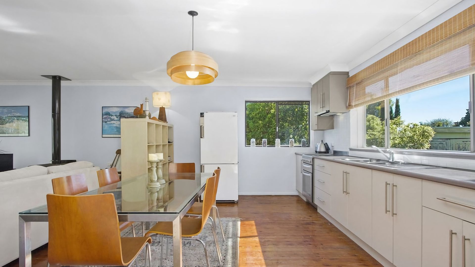 A modern kitchen with a dining area, featuring a glass table, wooden chairs, and a hanging light fixture.