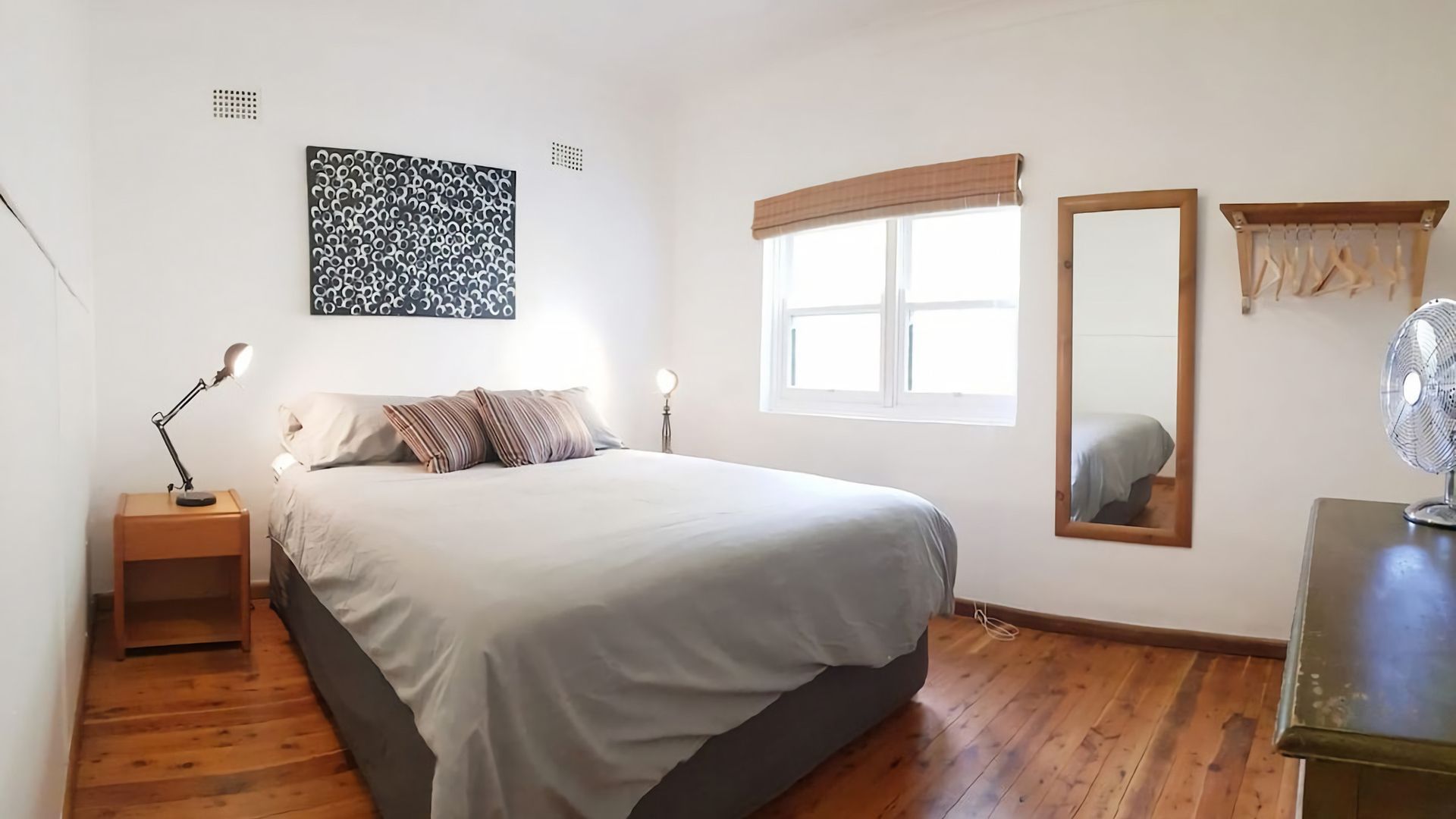 Une chambre à coucher avec un lit, des lampes de chevet, une fenêtre avec des stores, un miroir et un ventilateur sur pied.