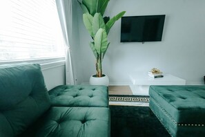 A modern living room with a green sofa, a potted plant, a coffee table, and a flat-screen TV mounted on the wall. - Extended-Stay 2BR · Monthly Rates · Near SFA (Nacogdoches)