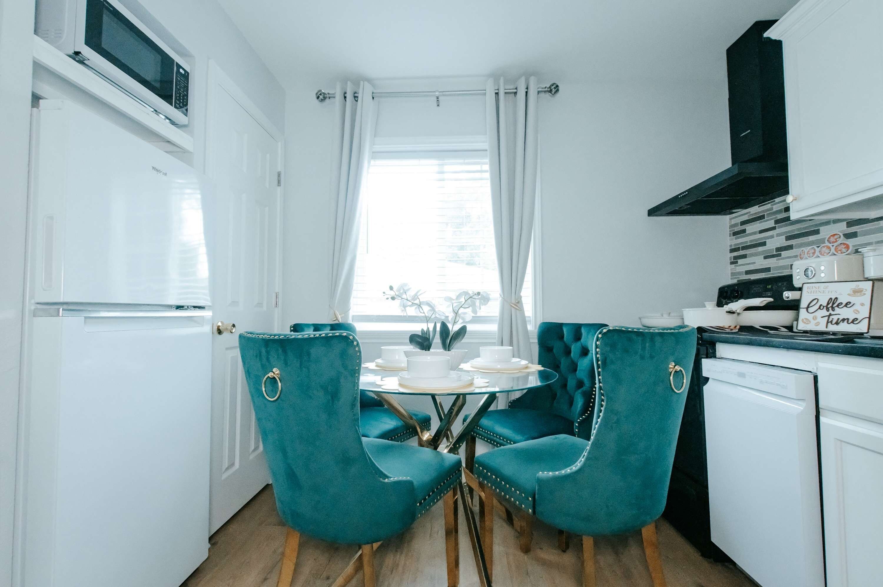 Une cuisine compacte avec des meubles blancs, un four à micro-ondes encastré, une cuisinière et un coin repas agrémenté de chaises turquoise et d’une table en verre.