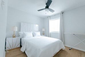 Une chambre à coucher comprenant un lit, deux tables de chevet, un ventilateur de plafond, une fenêtre avec des rideaux et un radiateur.