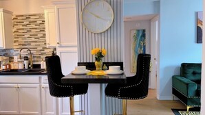 A kitchen with white cabinets, a black countertop, and a dining table with two chairs. There is a clock on the wall and a vase with flowers on the table.