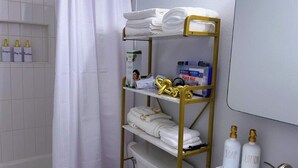 A bathroom with a white tiled wall, a shower curtain, a gold shelf with towels, and toiletries including soap and lotion bottles.