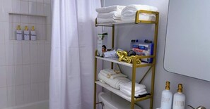 A bathroom with a white tiled wall, a shower curtain, a gold shelf with towels, and toiletries including soap and lotion bottles.