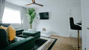 Ein modernes Wohnzimmer mit Deckenventilator, Flachbildfernseher, einem grünen Sofagarnitur, einer Topfpflanze und einem schwarzen Sessel.