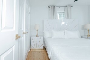 A white bedroom with a bed, bedside tables, and a window with curtains. - Extended-Stay 2BR · Monthly Rates · Near SFA (Nacogdoches)