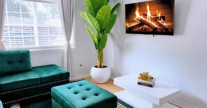 Un salon avec un canapé vert, une table d’appoint blanche, une plante en pot et une cheminée ornée d’un tableau encadré.