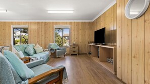 A room with a sofa, a TV on a wooden shelf, and a window with a view of trees. - Manyana Deck Life is a spacious pet friendly home (Manyana)