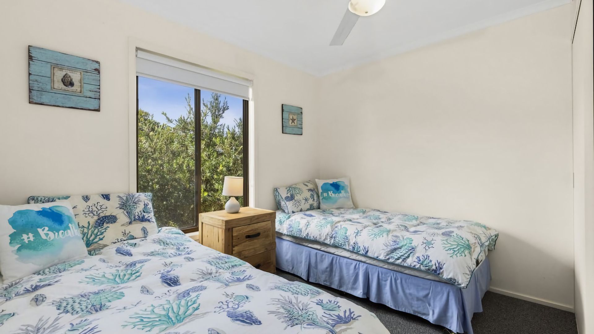 Ein Schlafzimmer mit zwei Betten, einem Nachttisch, einem Fenster mit Blick auf Grünflächen und einem Deckenventilator.