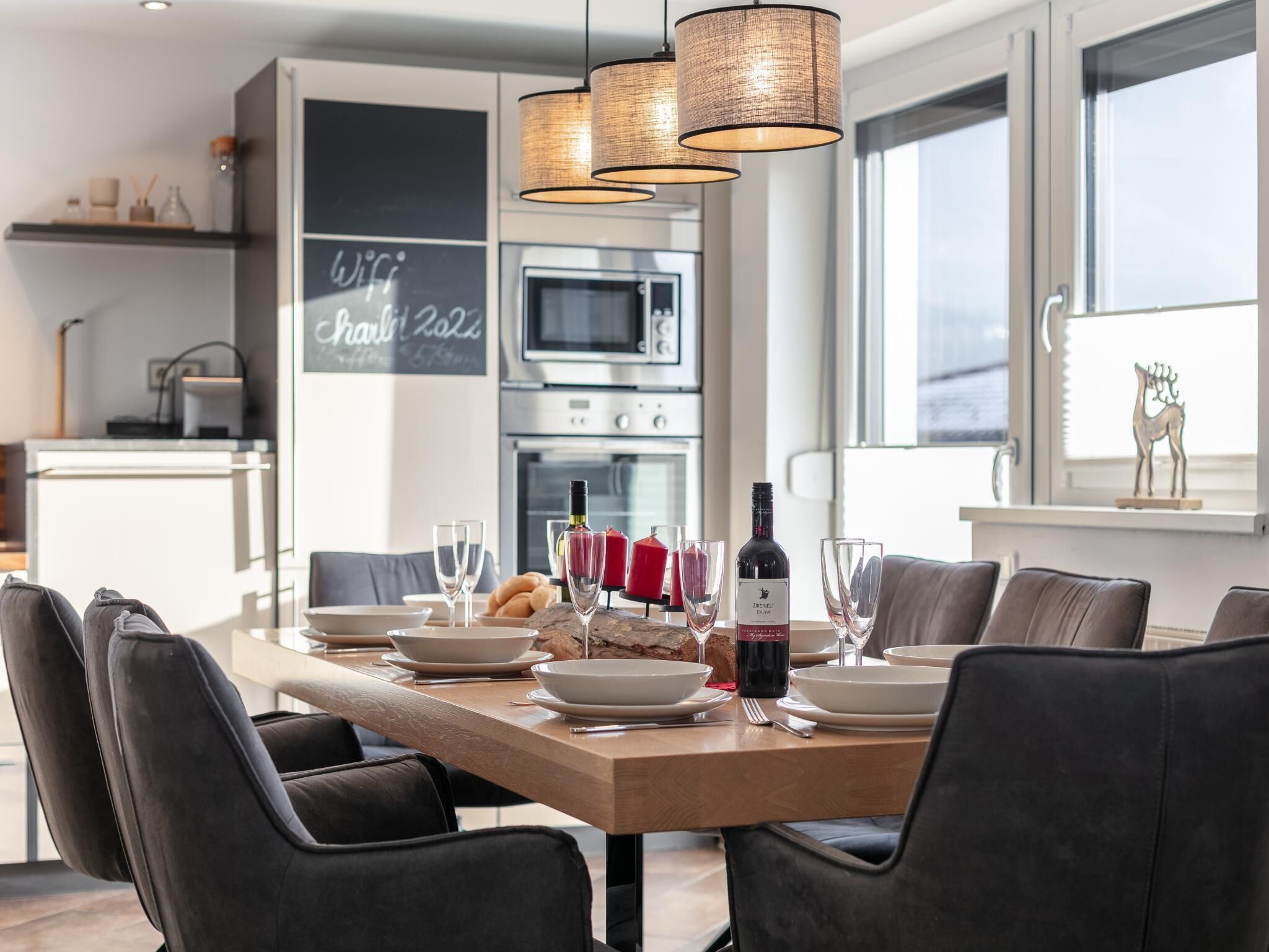 Un espace repas moderne avec une table en bois dressée pour six personnes, entourée de chaises sombres. Une bouteille de vin et des verres sont posés sur la table. La pièce est dotée d’une grande fenêtre, d’un tableau noir sur lequel est écrit le mot « WiFi », et d’une cuisine en arrière-plan.