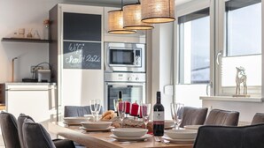 A modern dining area with a wooden table set for six, surrounded by dark chairs. A wine bottle and glasses are on the table. The room has a large window, a chalkboard with 'WiFi' written on it, and a kitchen in the background.