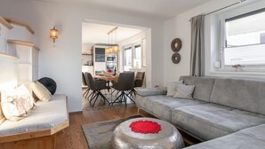 A modern living room with a grey sofa, a wooden bench, and a round silver table with a red centerpiece. There is a dining area with a table and chairs in the background.