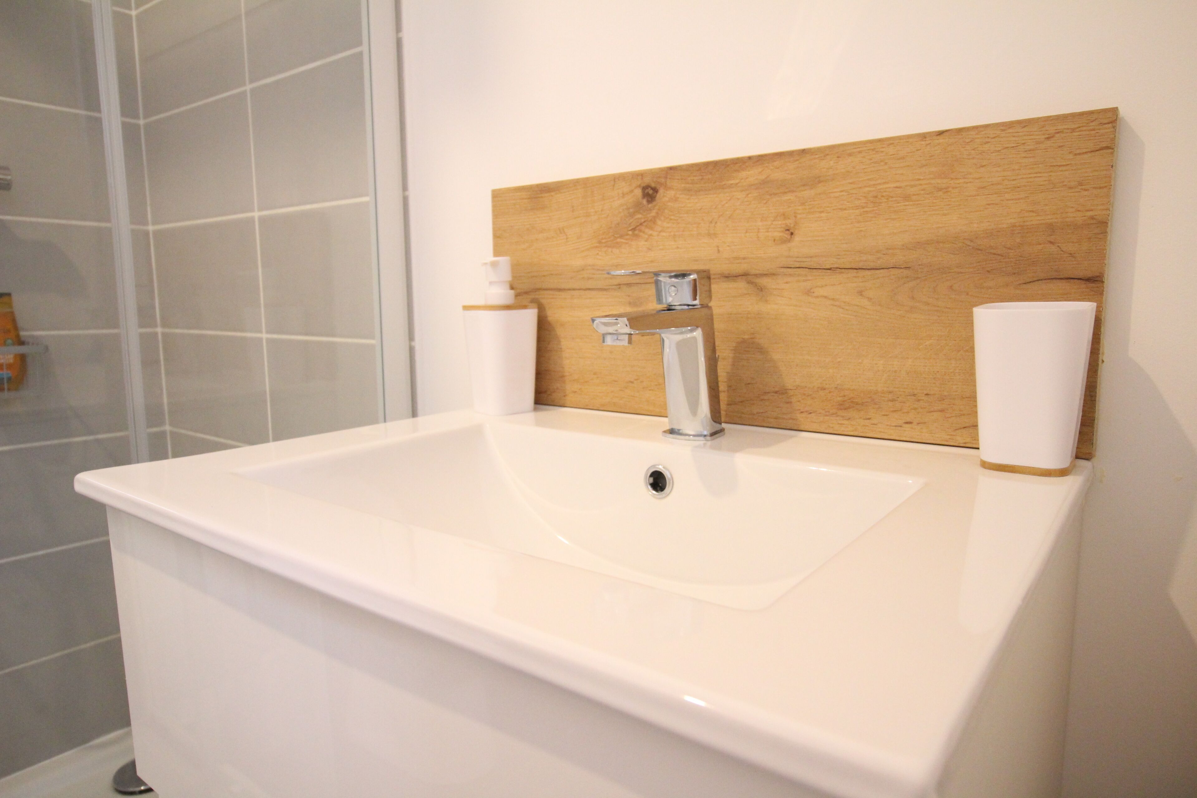 Un baño con lavamanos blanco, panel de madera sobre él y dos botellas blancas en la pared.