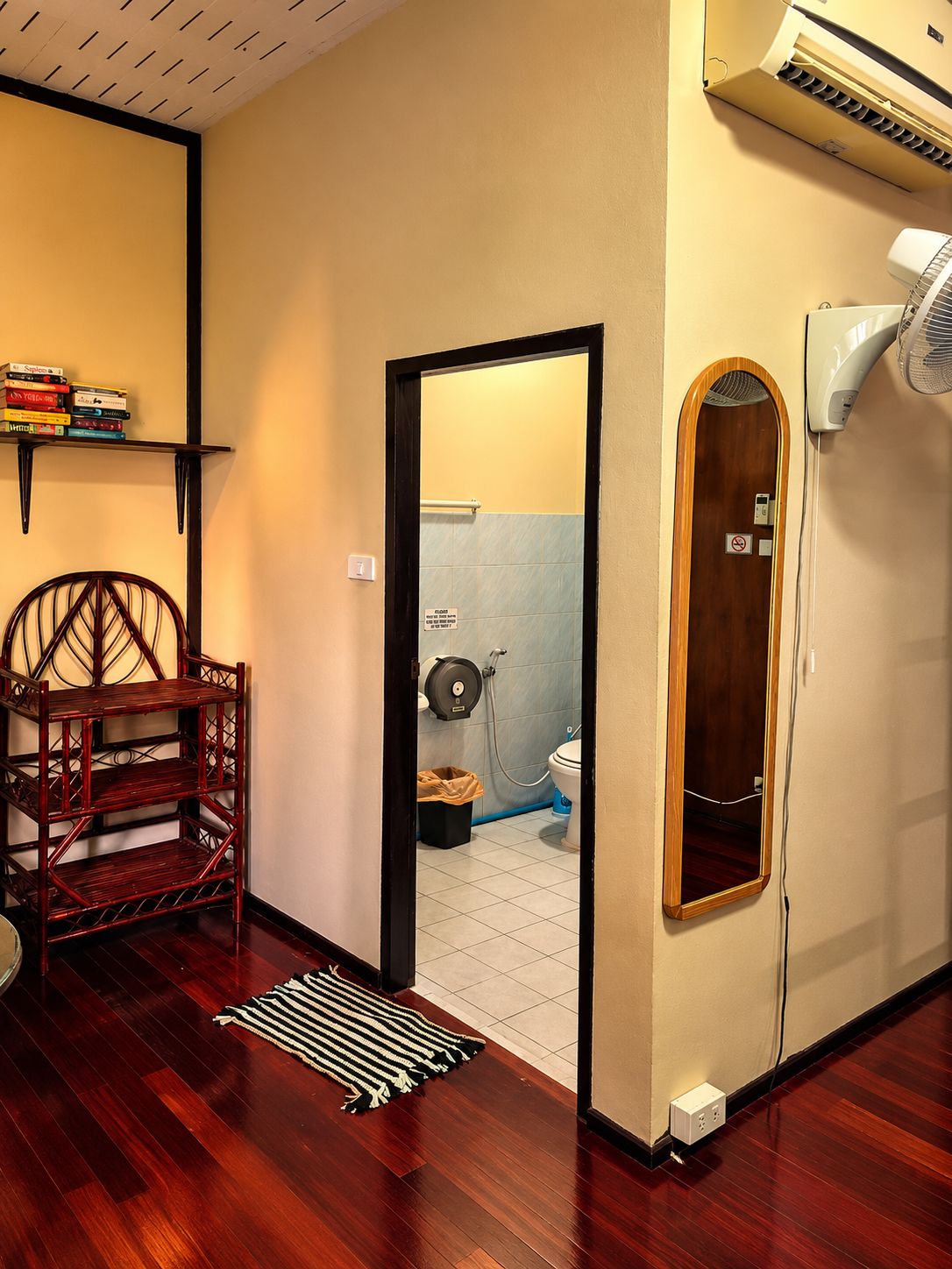 A room with a wooden chair, a small shelf with books, a door leading to a bathroom, and an air conditioning unit.