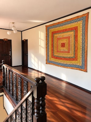 A spacious room with a wooden floor, a staircase with ornate balusters, and a large, colorful geometric patterned rug on the wall. - Marissa Hotel (Ko Pha-ngan)