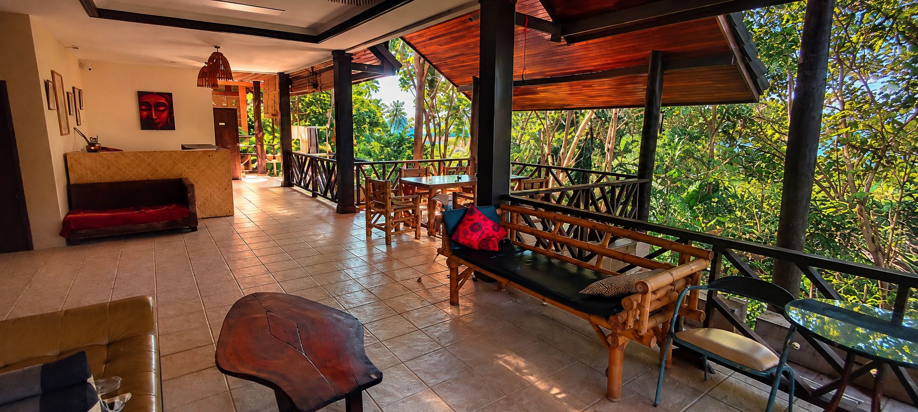A covered outdoor seating area with wooden furniture, a reception desk, and a view of the surrounding greenery.