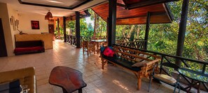 Área externa coberta com mobiliário de madeira, balcão de recepção e vista para o verde ao redor.