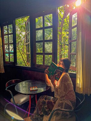 A person seated at a round table reading a book with a view of greenery through large windows. - Marissa Hotel (Ko Pha-ngan)