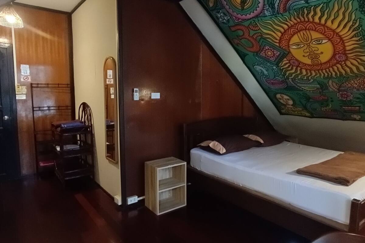 A bedroom with a wooden bed, a small wooden nightstand, a chair, a wall-mounted shelf, and a colorful tapestry.