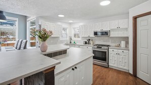 Une cuisine moderne avec des meubles blancs, un îlot central, des appareils électroménagers en acier inoxydable et une vue sur un paysage enneigé grâce à de grandes fenêtres.