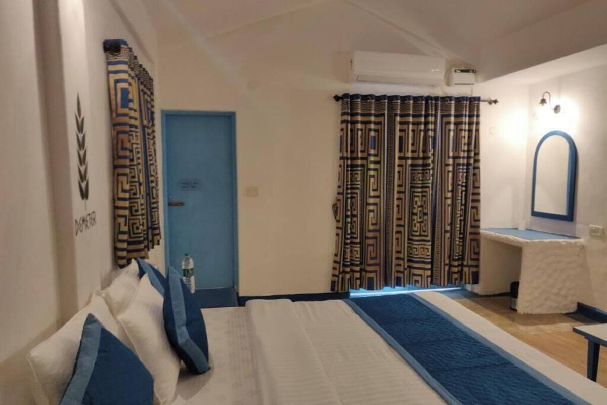 Un dormitorio con cama, una puerta azul, cortinas estampadas, una mesita blanca y un espejo.