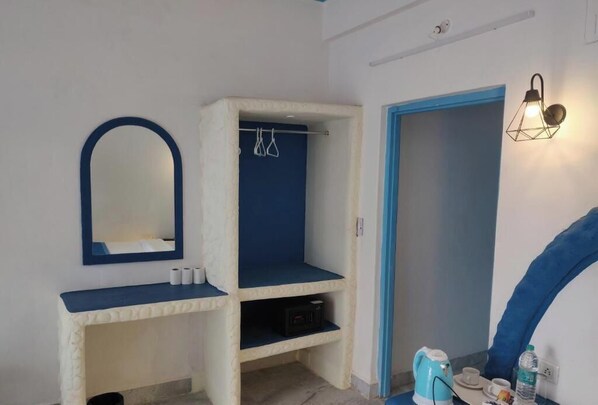 A bathroom with a mirror, a shelf with toiletries, a towel rack, and a wall-mounted light fixture. - Makarios boutique hotel  (Calangute)