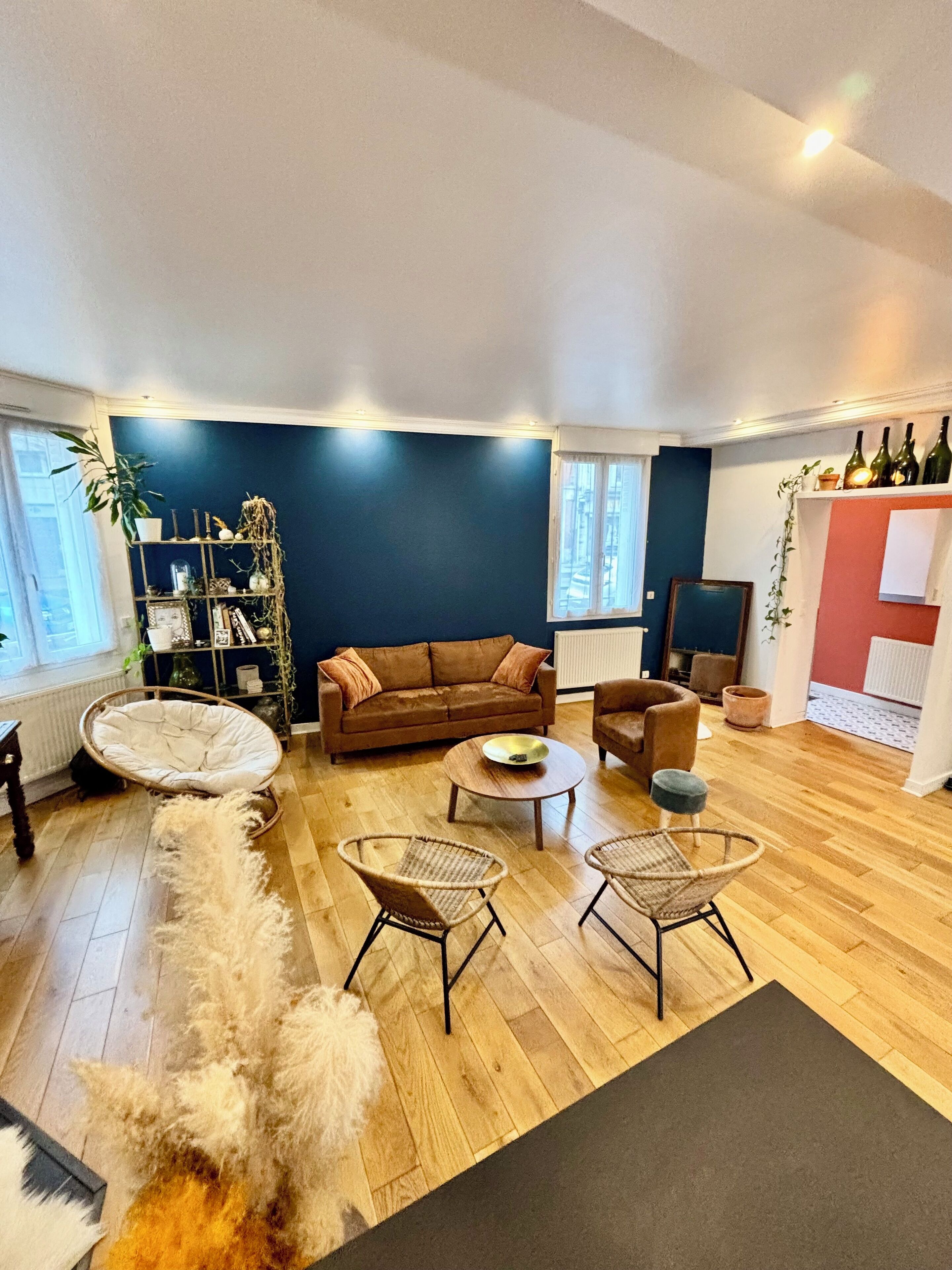 Un salon avec un canapé marron, deux fauteuils en osier, une table basse, un tapis moelleux et une bibliothèque.