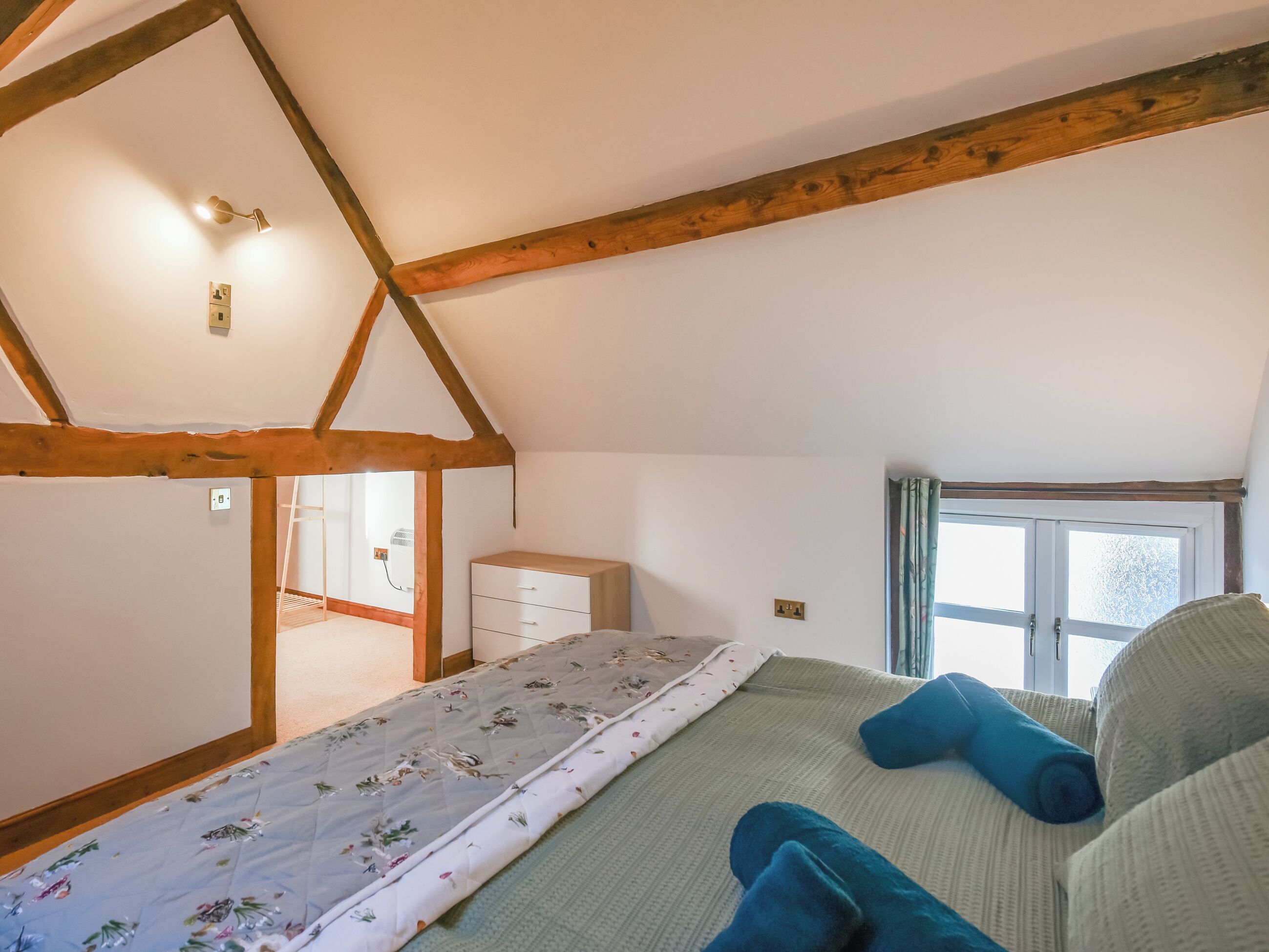 A bedroom with a sloped ceiling, a bed with floral bedding, a dresser, and a window with curtains.