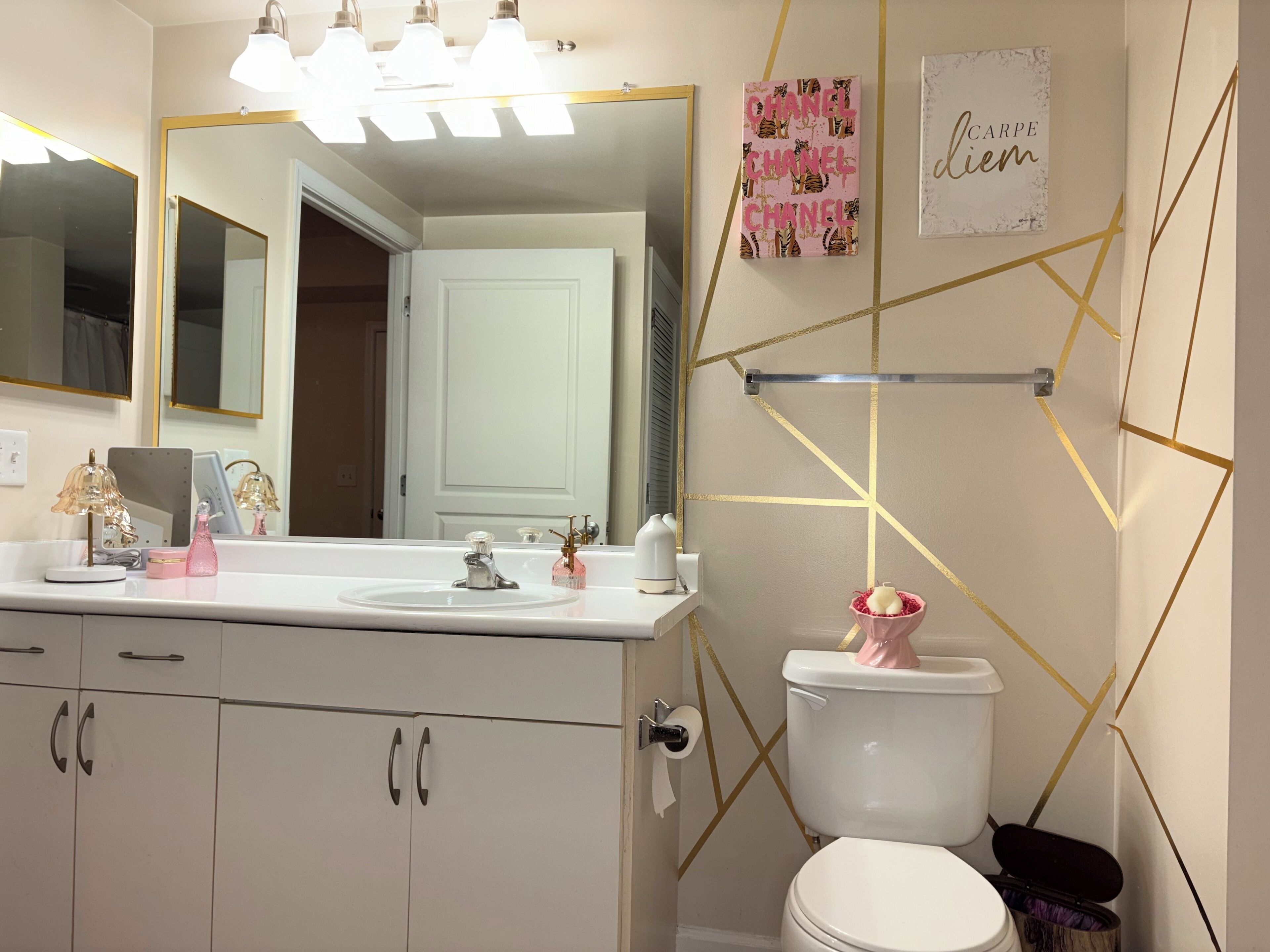 Une salle de bain avec un lavabo blanc, un miroir et des toilettes. On trouve des objets décoratifs sur le lavabo et un porte-serviettes au-dessus des toilettes.