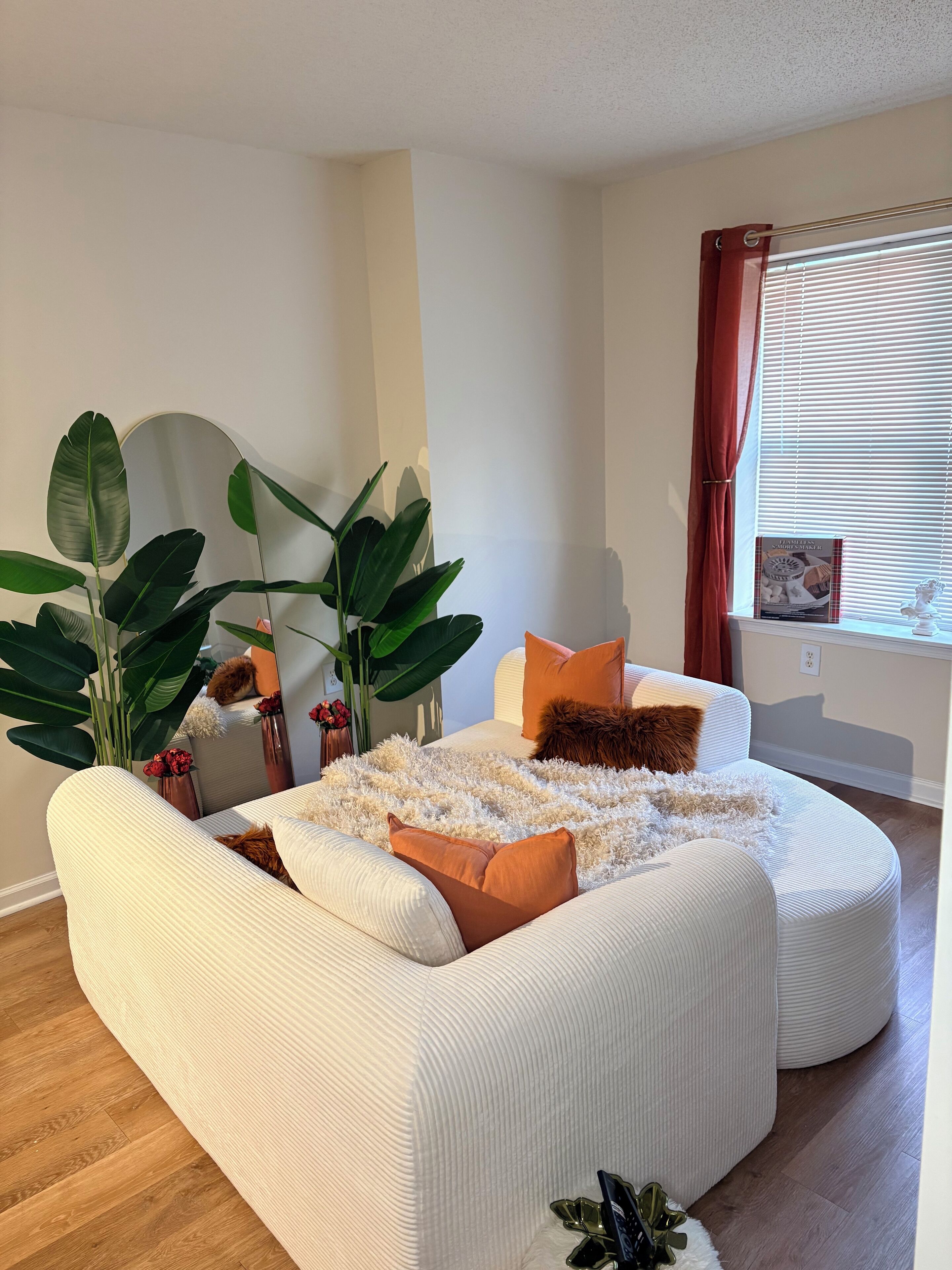 A modern living room with a white sofa, large green plants, and a window with blinds.