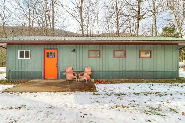 A small, single-story building with a green exterior and an orange door. There are two Adirondack chairs outside, and the ground is partially covered with snow.